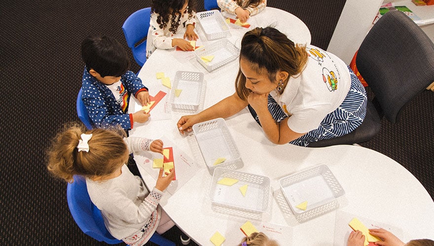 Definition of Education, featuring a teacher observing 6 young kids in a Shichida class. The kids are solving a 3d tangram puzzle challenge.
