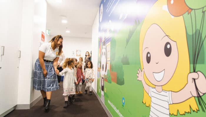 A Shichida teacher, happily leading 6 preschool kids to their classroom. They are walking past a colourful wall mural.