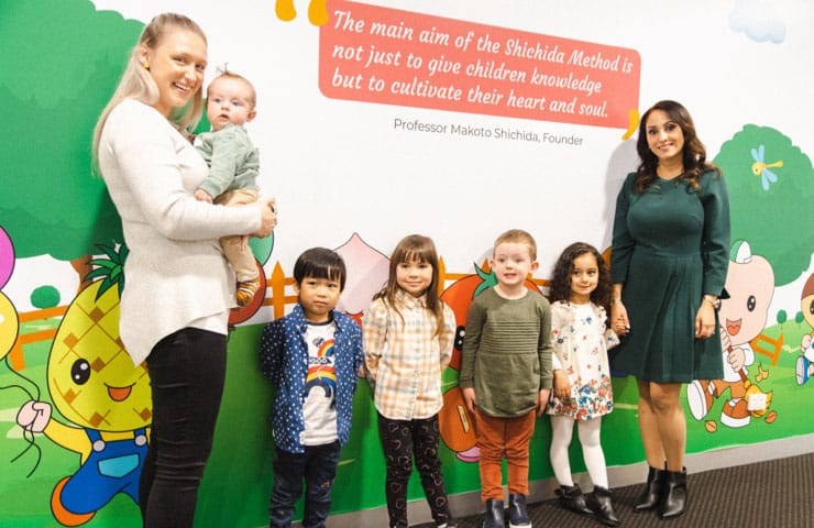 why shichida early learning centre? Featuring two mums, 4 preschoolers and a baby. They are standing in front of a colkourful mural in a Shichida centre