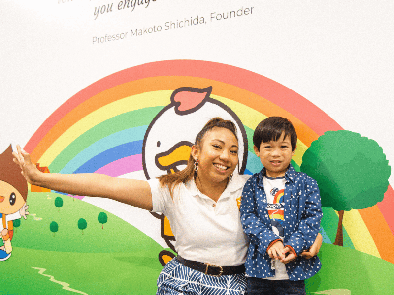 Shichida Promotions - A friendly Shichida teacher smiles alongside a young child. They are in front of a colourful mural featuring a rainbow.