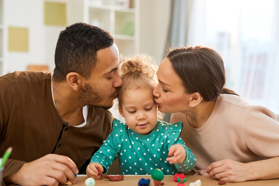 Kids mental health, featuring a mum and dad kissing their young daughter on the head, while she plays with toys.