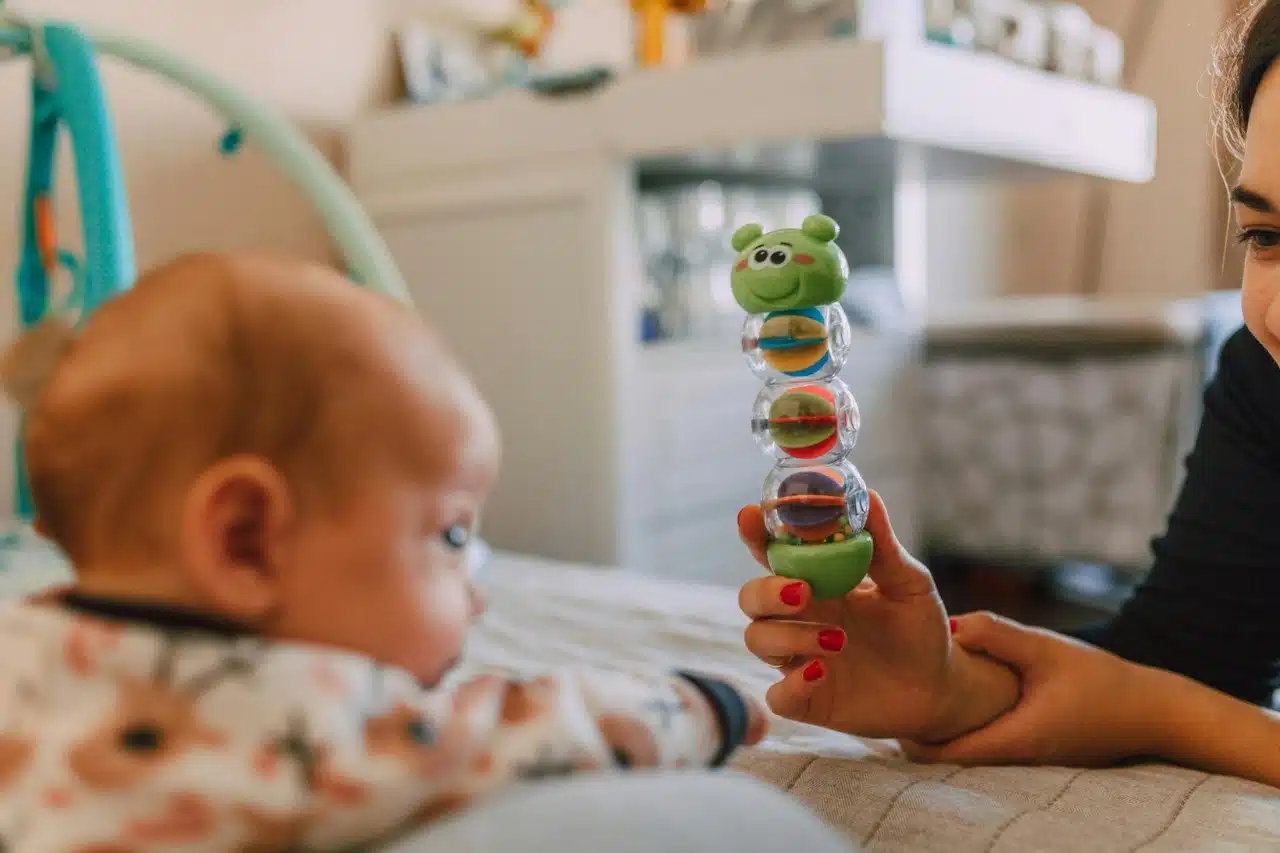 A woman holds a colorful toy in front of a baby, who looks curiously at the toy