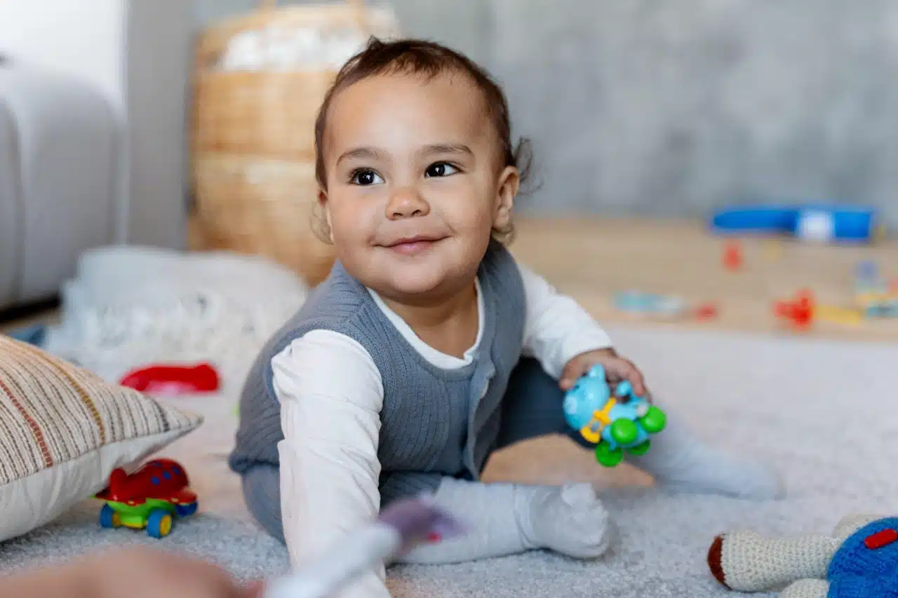 A baby sits on the floor, happily playing with colorful toys scattered around