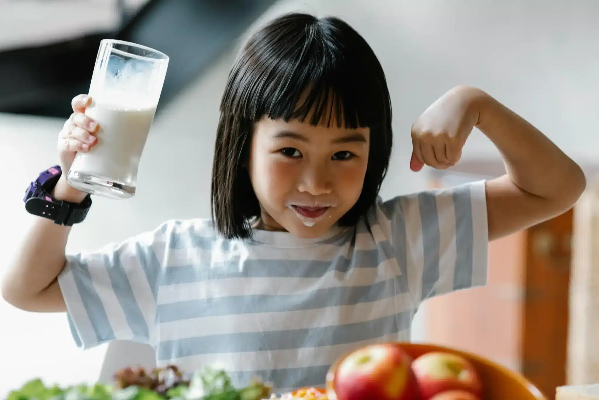A young girl with milk on her face, holding a glass of milk and flexing her arm with a healthy meal in front of her.