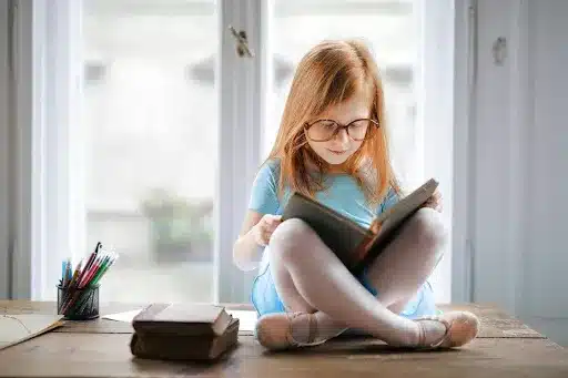 How to Improve Concentration in Kids - Young girl with glasses sitting cross-legged on a table, reading a book near a bright window.