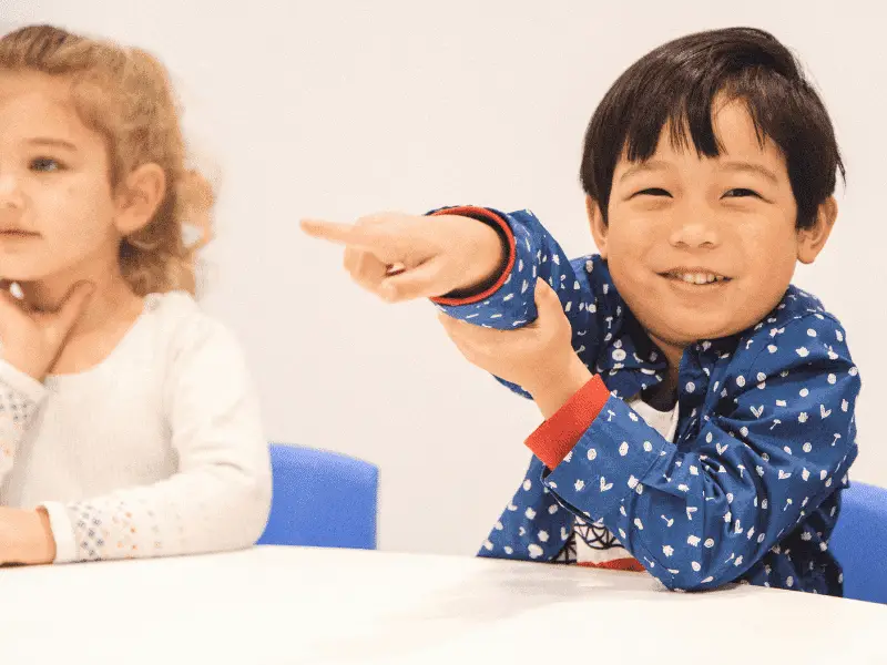 Smiling young boy pointing playfully while sitting next to a girl in a classroom setting.