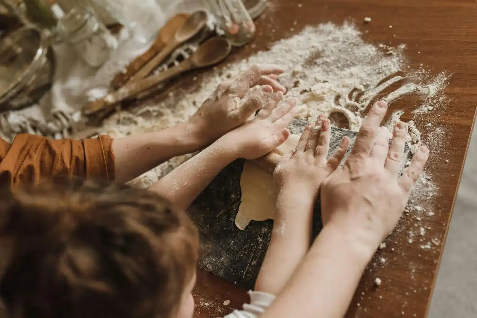 An overhead view of an adult's hands gently guiding a child's hands as they press down on a piece of dough on a flour-dusted wooden table.