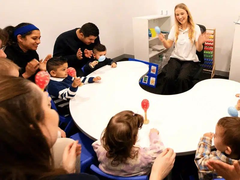 A female teacher leads a parent-child music class, sitting at the head of a white table and shaking blue maracas while smiling at the toddlers and adults seated around the table.