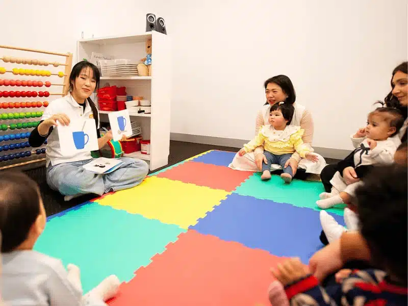 A female teacher sits on a colorful playmat holding up a flashcard of a blue cup, leading a lesson for a group of parents and toddlers in a classroom.