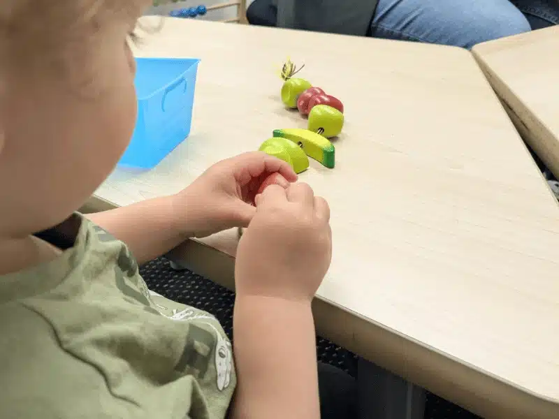 A close-up over-the-shoulder view of a toddler in a green shirt sitting at a wooden table, playing with a line of small, wooden toy fruits. Sensory Activities for Toddlers