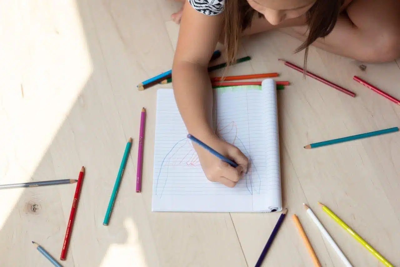 Photographic memory in kids - A child draws in a notebook on the floor, surrounded by colorful pencils.
