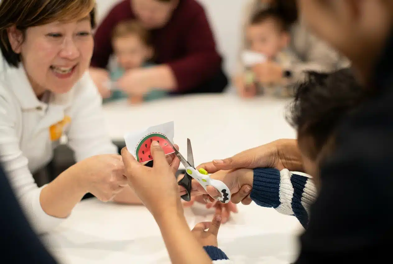 instructor guides a preschooler while cutting a rounded shape with safety scissors