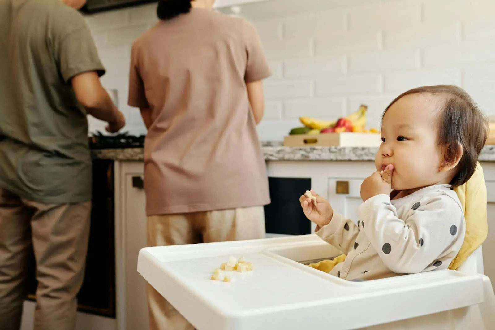 Toddler in a high chair eating small pieces of food while two adults prepare a meal in a kitchen background - pincer grasp