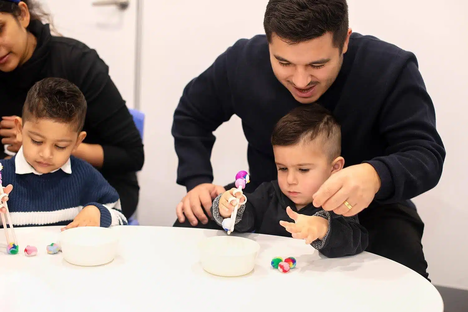 A young boy practices fine motor skills using training chopsticks with his father's guidance during an educational activity.