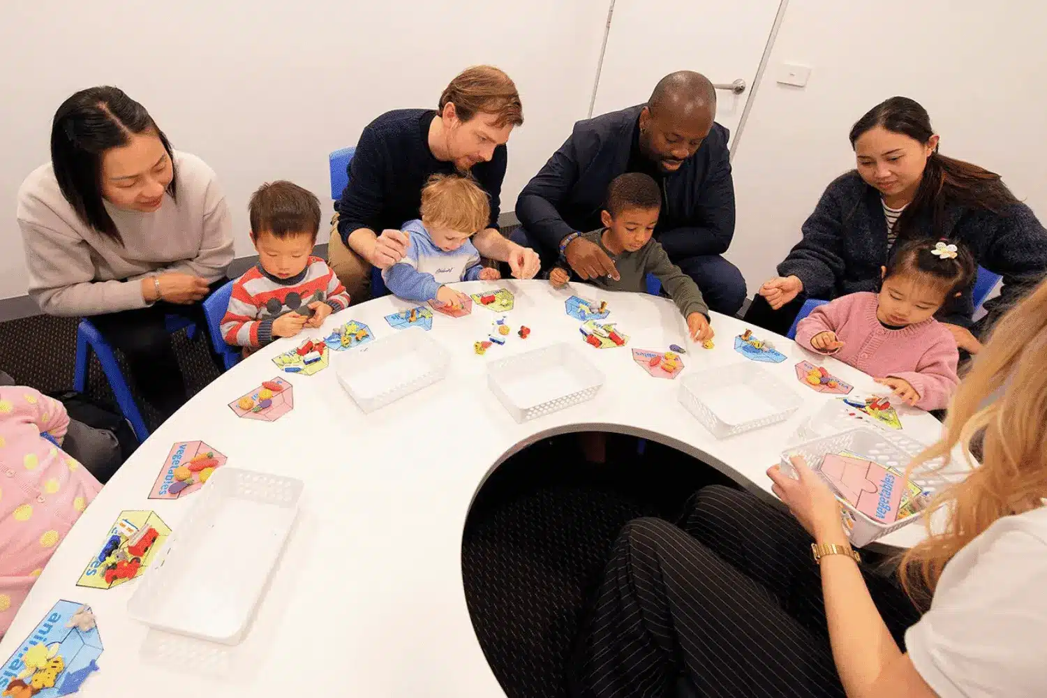 Diverse group of parents and young children sitting around a white table participating in an educational sorting activity.