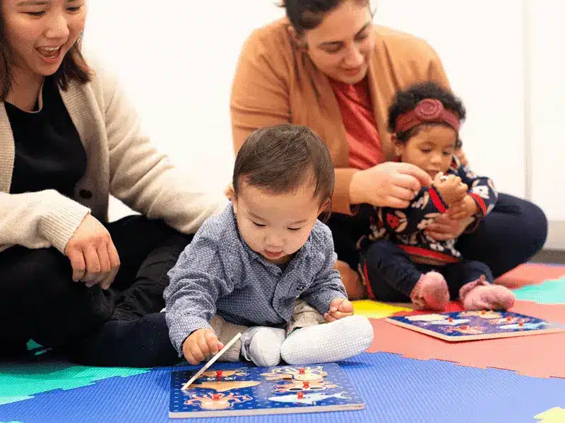 Two women supervising a young boy and girl as they play with sea-animal themed wooden puzzles on a colorful foam play mat.